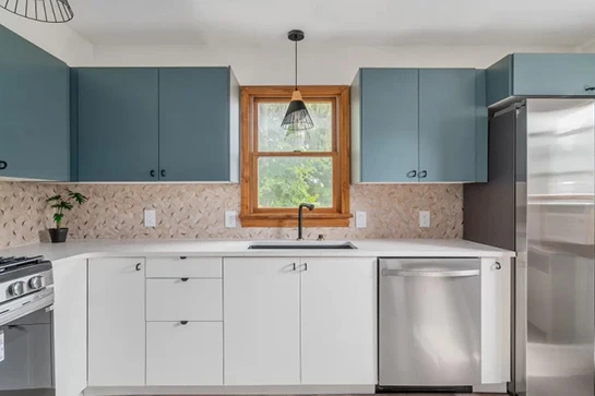Modern kitchen with blue upper cabinets, white lower cabinets, stainless steel appliances, and a wood-framed window.
