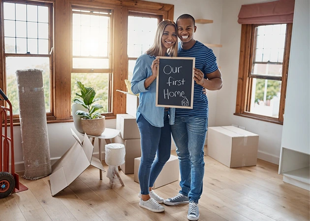 Happy couple in new home, holding "Our first home" sign. First-time buyer property showing.