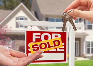 House keys being handed over in front of a "For Sale" sign marked "Sold.